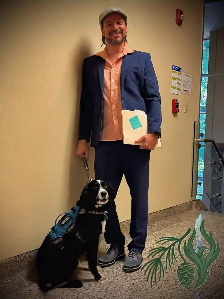 Kyle McCall, a corporate clinical consultant wearing a blue suit jacket and holding a manila folder, stands next to his black and white service dog in an office hallway.